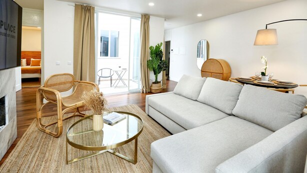 A modern living room with a sofa, a glass coffee table, and a wicker chair. There is a dining table with a lamp and a potted plant. - Green Algarve Gem Spacious 2 BDR By Misha s Place (Faro)