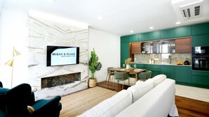 Ein modernes Wohnzimmer mit einem weißen Sofa, einem Essbereich mit Holztisch und -stühlen, einem an einer Marmorwand montierten Fernseher und einer Küche mit grünen Schränken und einem schwarzen Backofen.