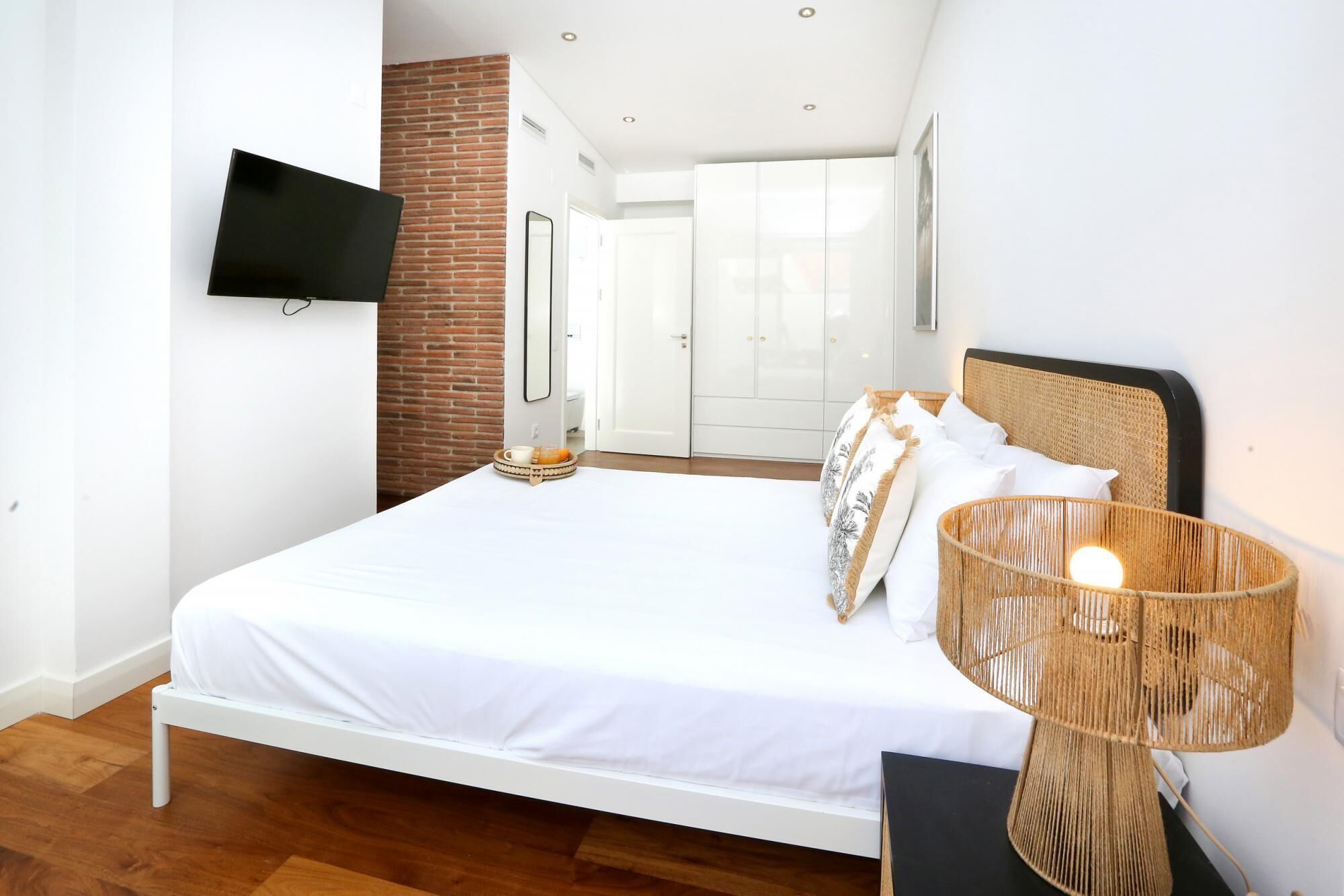 Un dormitorio con una cama blanca, un televisor de pantalla plana montado en la pared, una lámpara de mimbre y una pared de ladrillo decorativa.