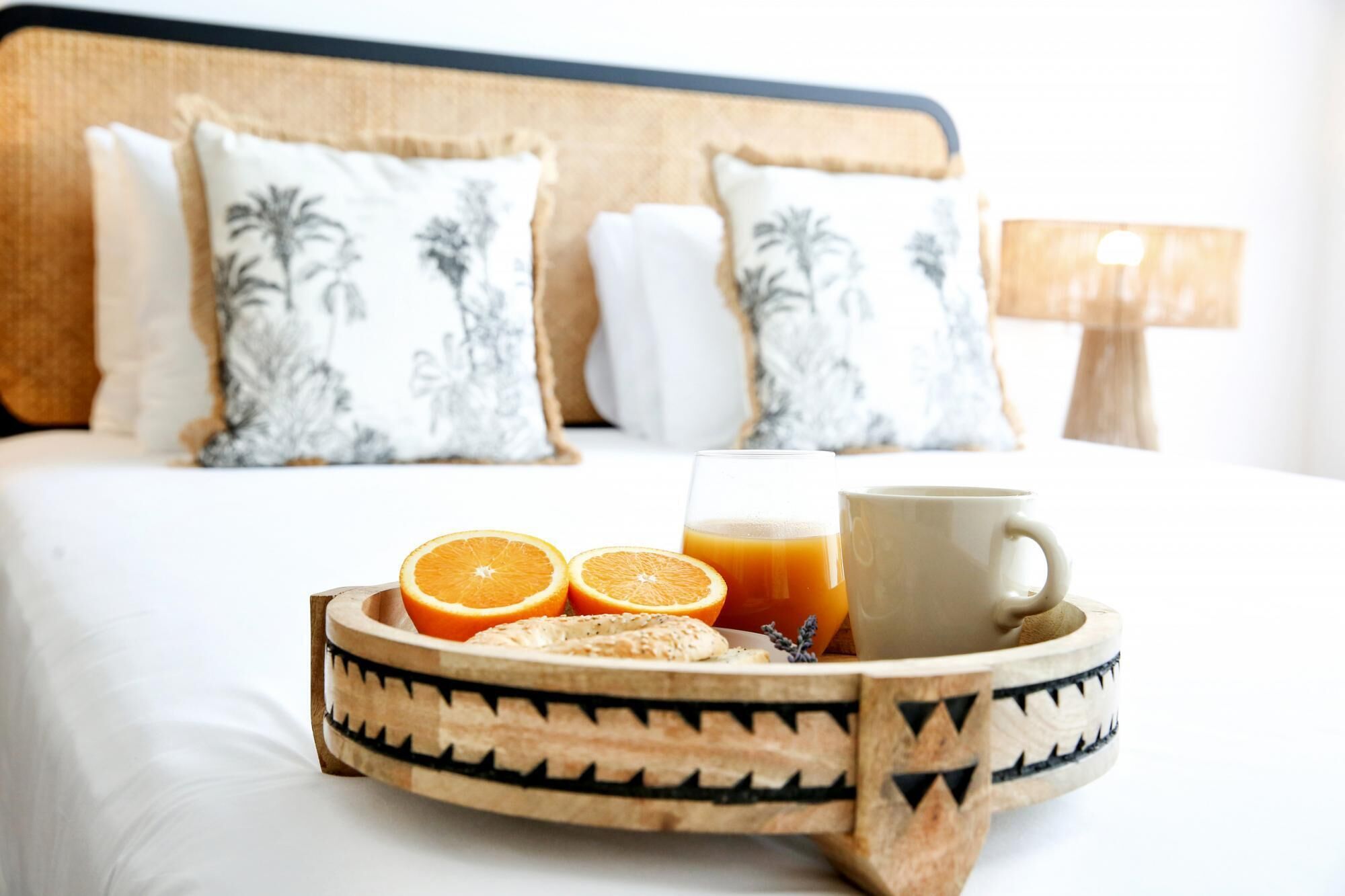 A neatly made breakfast tray with orange slices, a glass of orange juice, and a cup on a bed with a wooden headboard and patterned pillows.
