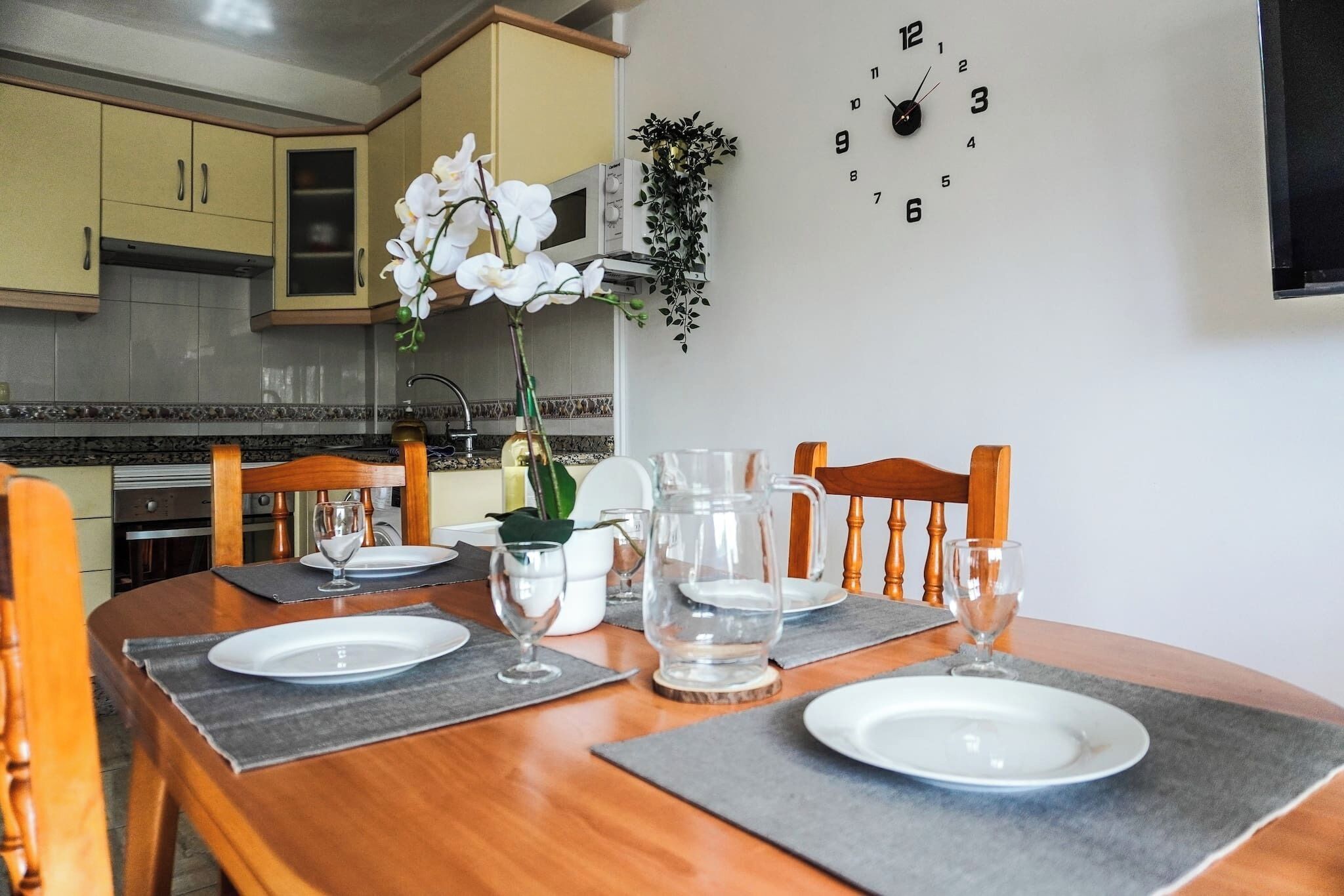 Une table à manger est dressée avec des assiettes, des verres et un pichet, entourée de chaises, dans une cuisine dotée de placards et d’une horloge au mur.