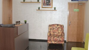 Un área de recepción con un escritorio de madera, un sillón floral, una silla verde, una araña de techo, un cuadro enmarcado, una bandera roja y un televisor en la pared.