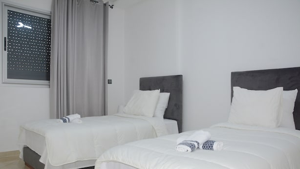 Dos camas individuales con ropa blanca y cabeceras, una ventana con vista al cielo y una cortina medio recogida.