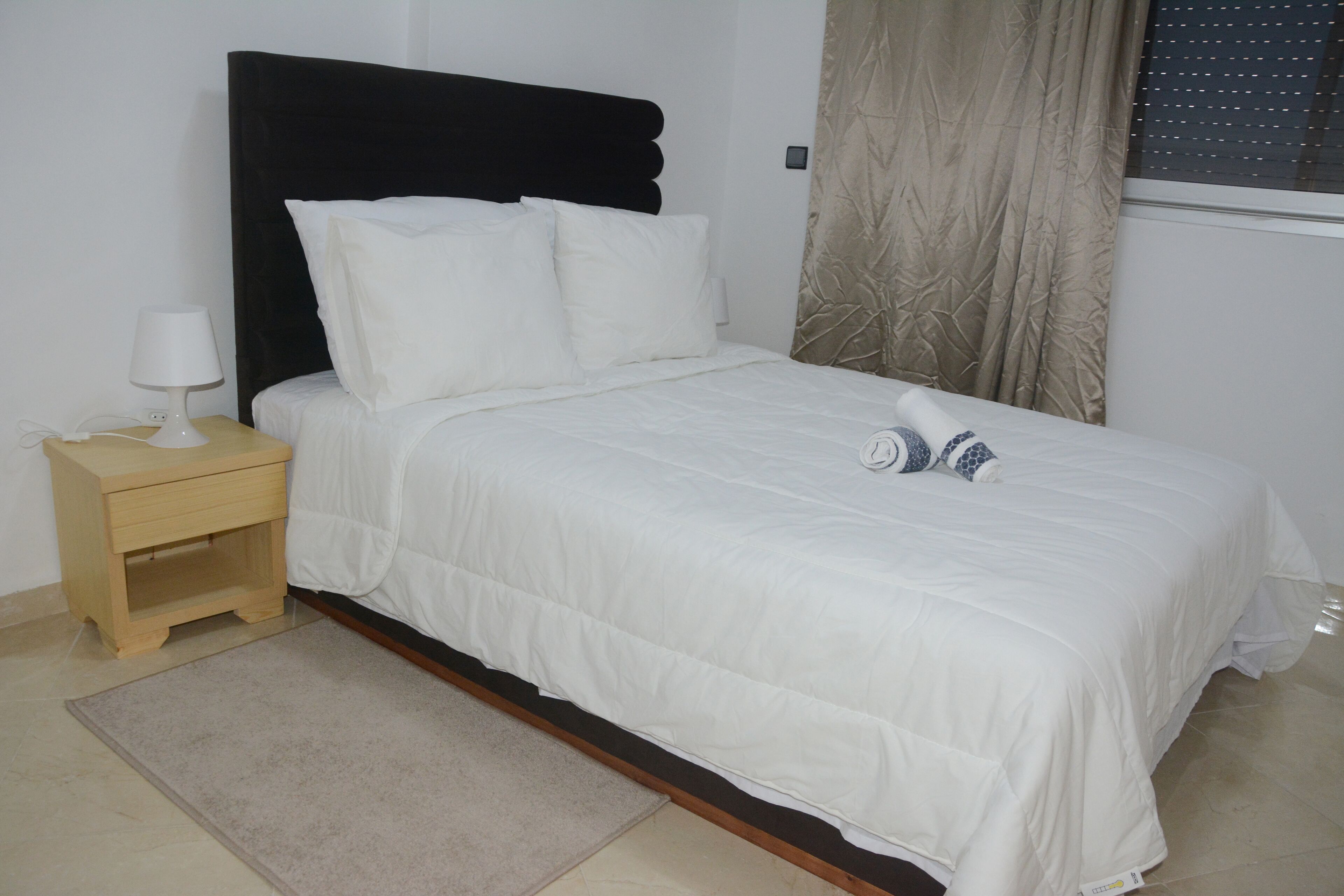 A neatly made bed with white bedding, a wooden nightstand with a lamp, and a beige rug.