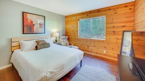 Une chambre avec un plafond en bois, un lit avec une literie blanche, une table de chevet avec une lampe, une chaise et une fenêtre donnant sur des arbres.