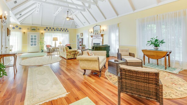 A spacious living room with hardwood floors, a high ceiling with exposed beams, and a variety of seating including a sofa, chairs, and a wicker armchair.