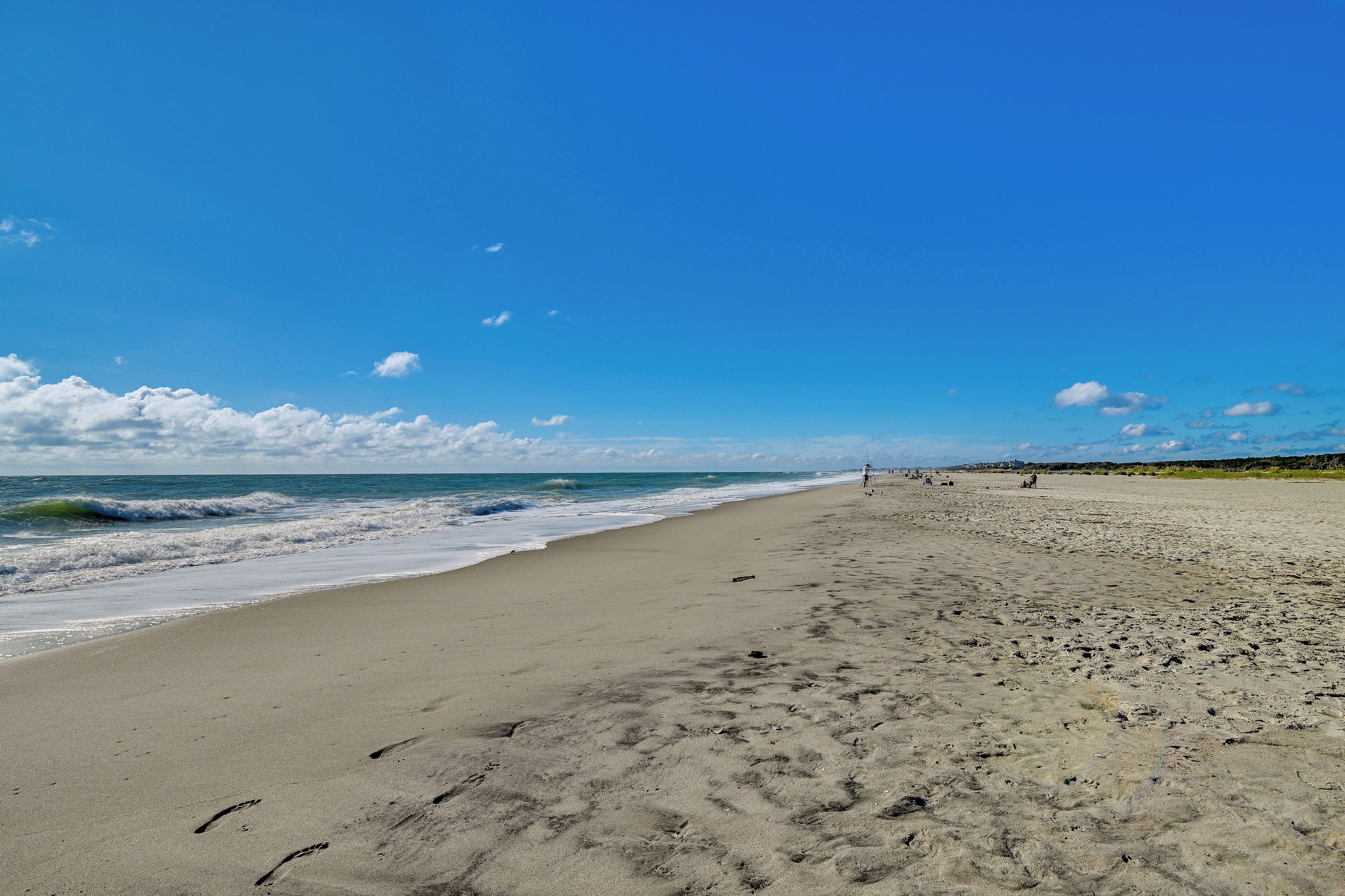 A wide sandy beach with footprints leading to the water, a clear sky, and distant hills.