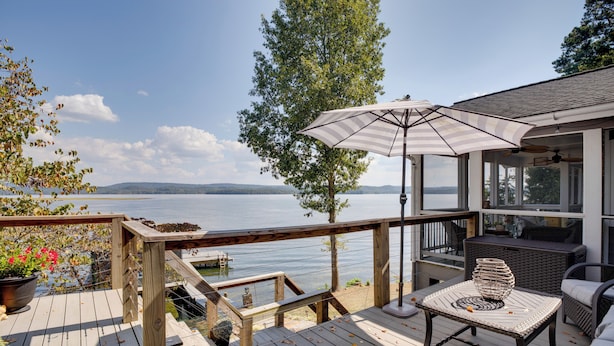 Une terrasse en bois avec des meubles d’extérieur, un grand parasol et une vue sur un lac.