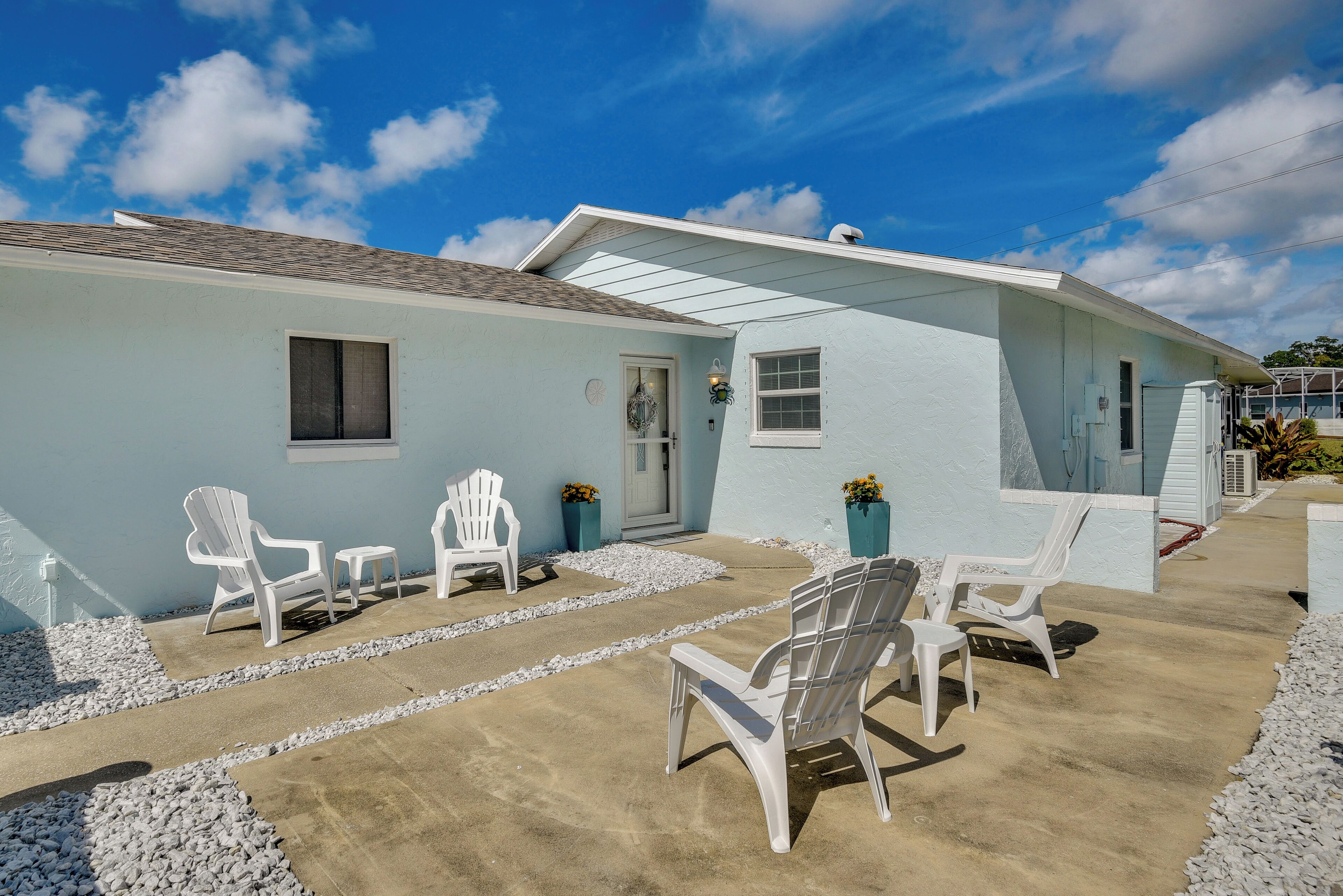 A light blue house with a covered patio area featuring white Adirondack chairs and a concrete pathway.