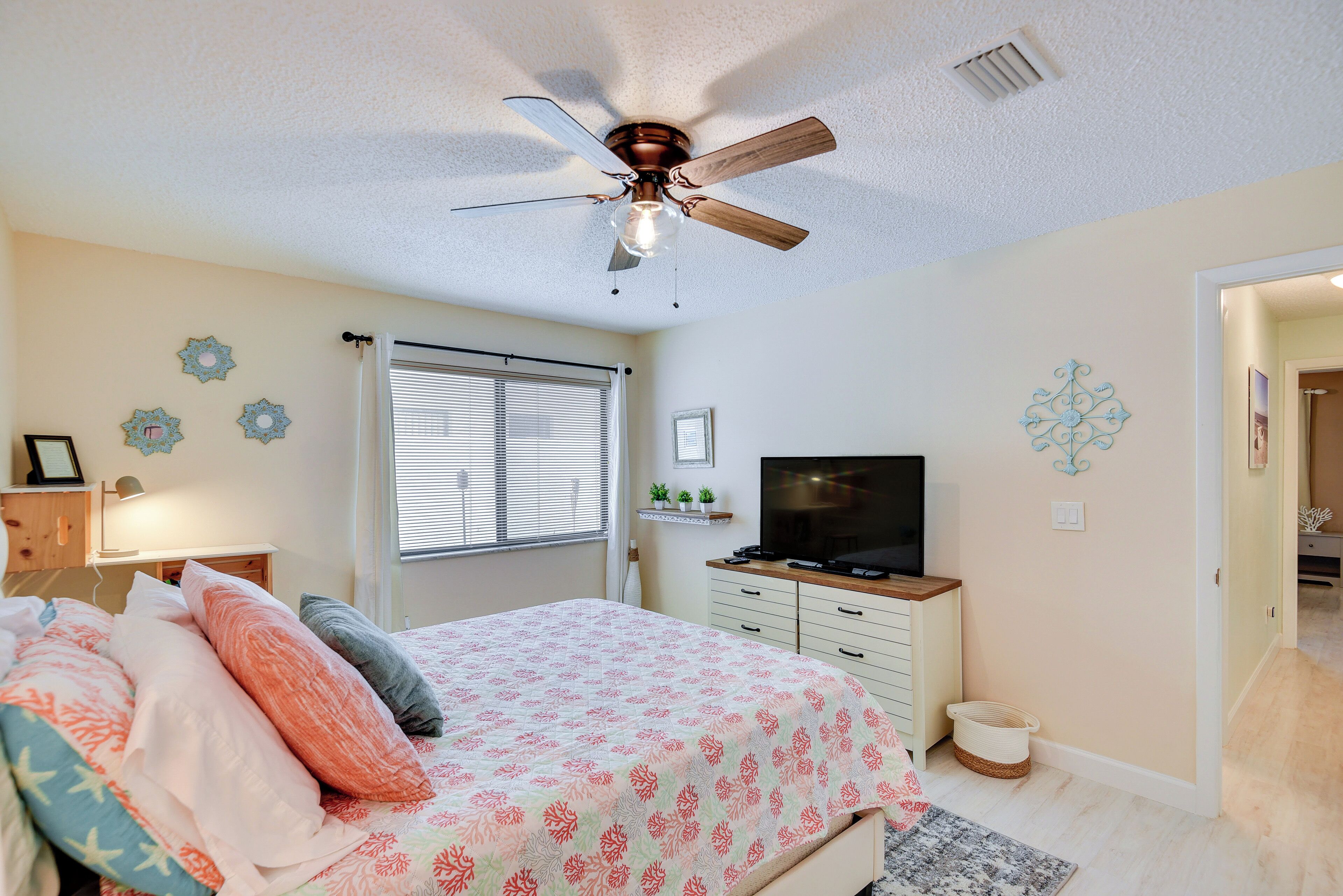 Une chambre à coucher comprenant un lit, un ventilateur de plafond, une télévision, une commode et des œuvres d’art décoratives au mur.