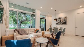 A modern living room with a large window, a sofa, a coffee table, and a dining area with chairs. - Custom New Build Central Austin Home Mins to DT (Austin)