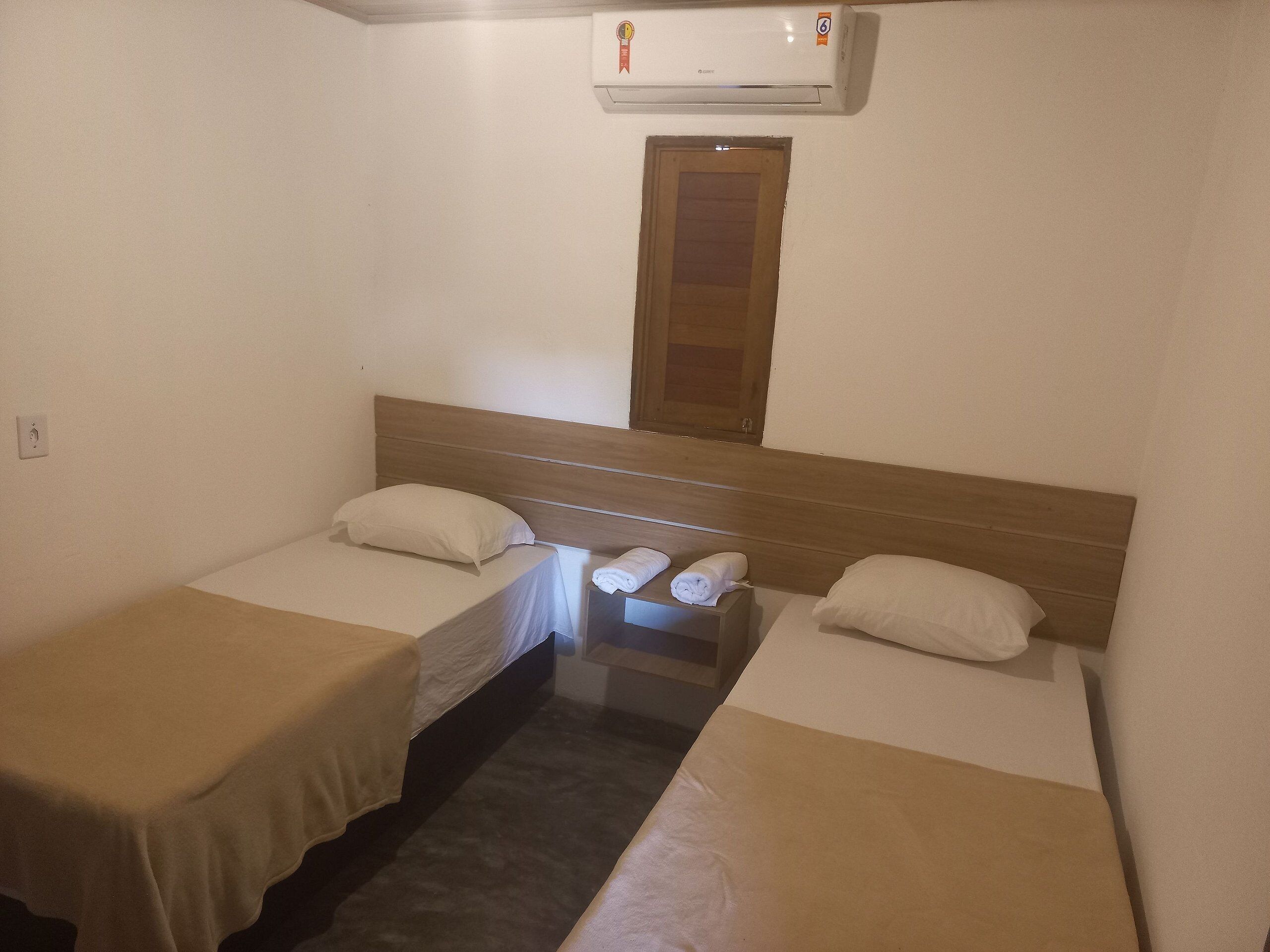 Una habitación pequeña y limpia con dos camas individuales, cabecera de madera, una mesita con toallas y un equipo de aire acondicionado sobre la puerta.