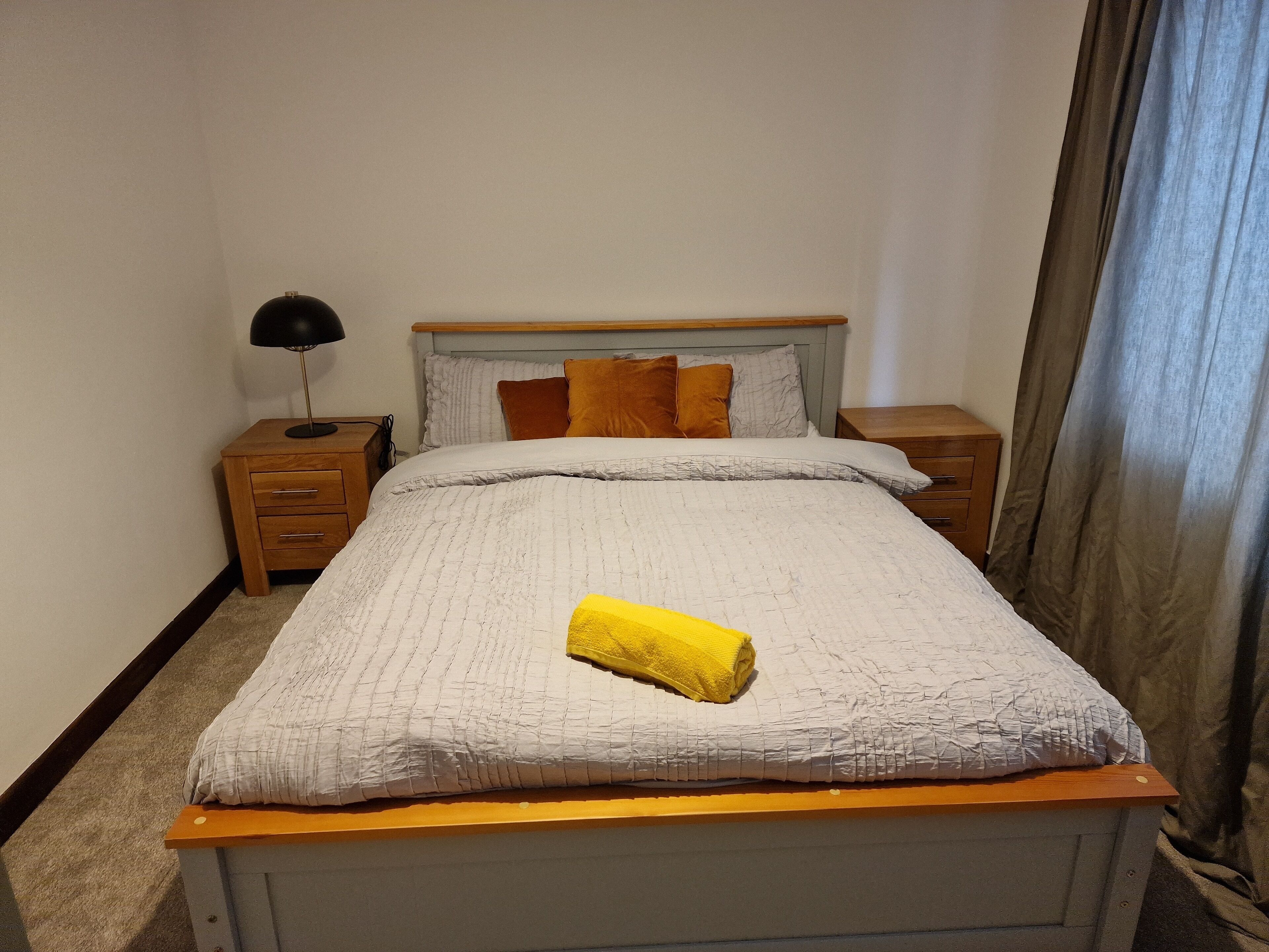 Una cama bien hecha con colcha blanca, cabecera de madera y toalla amarilla sobre la cama. Hay dos mesitas de noche con lámparas, y una cortina marrón en la ventana.