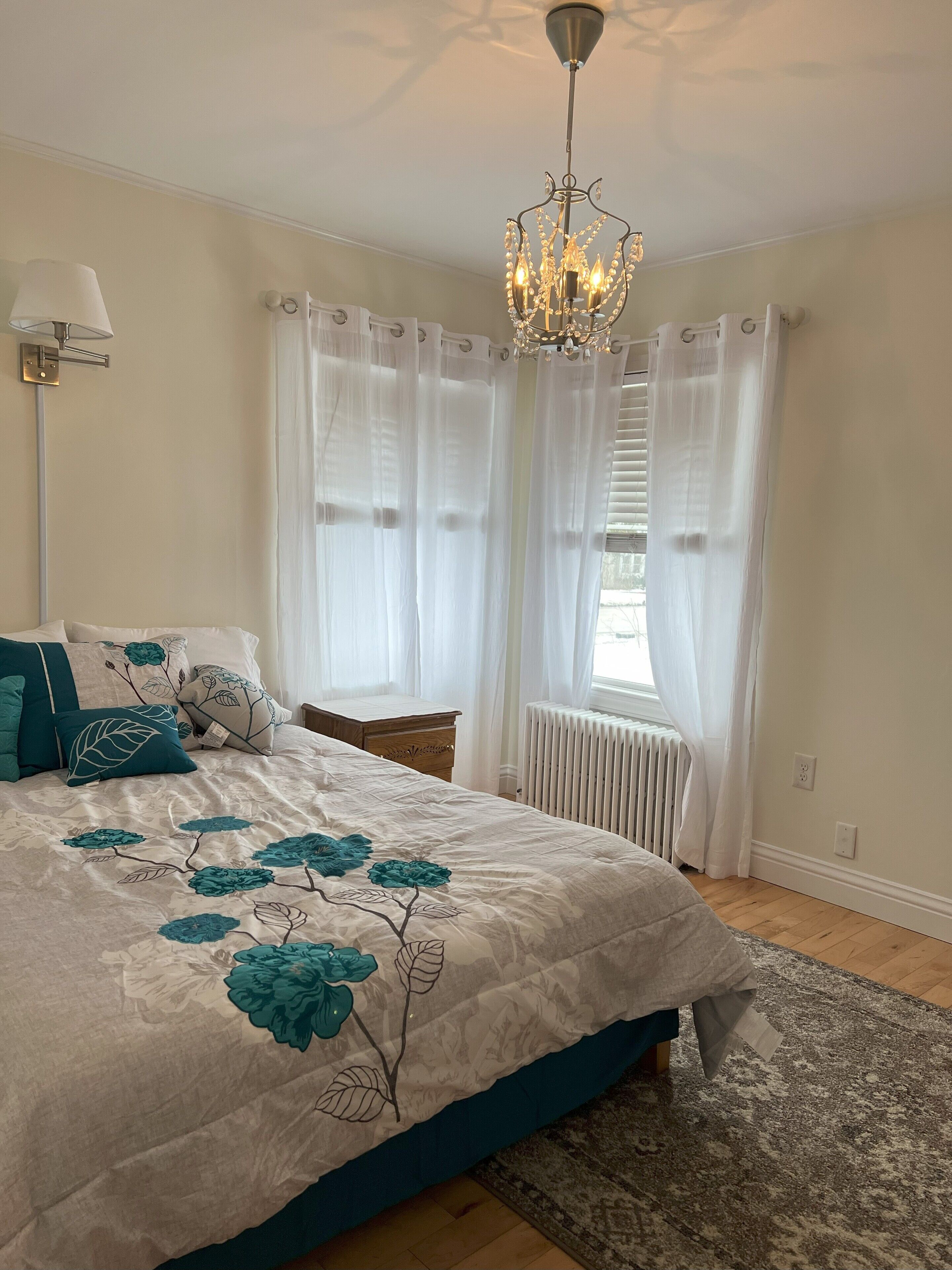 A bedroom with a large bed, a chandelier, white curtains, a radiator, and a nightstand.