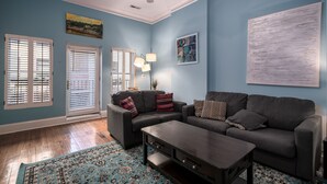 Una sala de estar con pared azul, dos ventanas con persianas, un sofá gris, una mesa de centro y un tapete estampado.