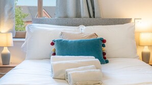 Uma cama bem arrumada com lençóis brancos, um travesseiro azul com pompons coloridos e um cabeceira cinza.