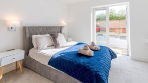 Quarto moderno com cabeceira estofada cinza, cama grande com colcha azul, duas luminárias de cabeceira e vista para um carro vermelho pela janela.