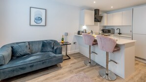 A modern living room with a grey sofa, a small side table, and a kitchen area with a microwave, toaster, and bar stools.