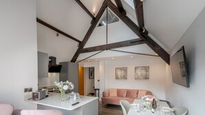 Et moderne køkken med en central ø, et spiseområde med et hvidt bord dækket til to, og en hyggelig opholdsstue med en sofa.
