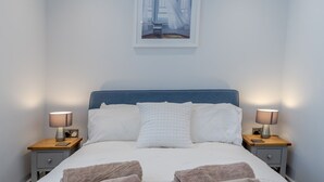 En pænt redt seng med hvide sengetøj og to puder, med to sengeborde med lamper på hver side, i et rum med et enkelt, rent design.