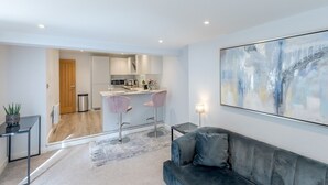Una sala moderna con un sofá gris, una zona de cocina con armarios blancos y un cuadro abstracto de gran tamaño en la pared.