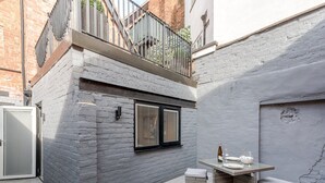 Un pequeño patio exterior con una mesa y dos sillas de mimbre, una ventana y un balcón con barandillas de metal.