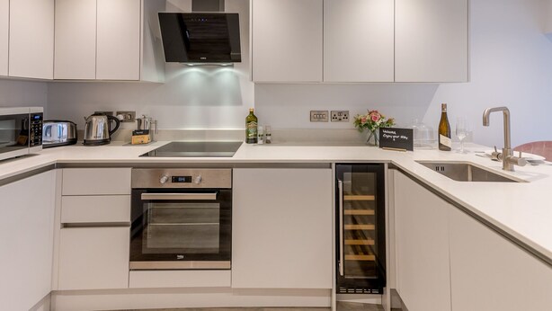 Una cocina moderna con muebles blancos, horno integrado, microondas, cafetera y una botella de vino.