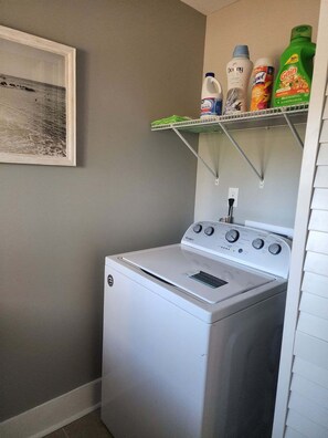 A laundry area with a front-loading washer and dryer, a shelf with cleaning supplies, and a framed picture on the wall.
