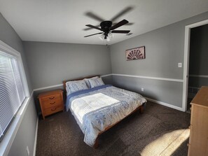 Quarto com uma cama de solteiro, um ventilador de teto, uma mesinha de cabeceira e uma janela com persianas.