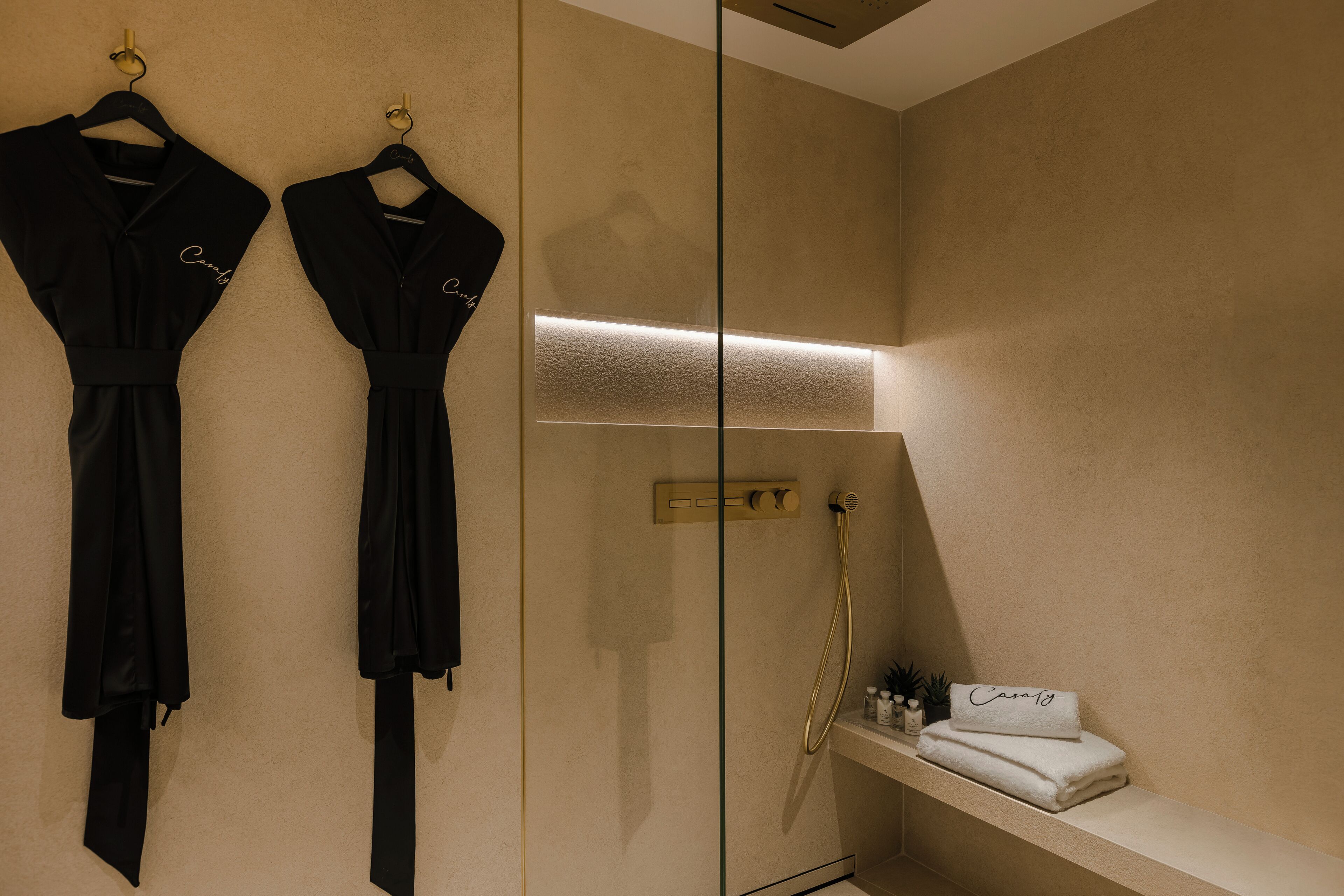 Un baño con dos batas negras colgadas en ganchos, una cabina de ducha de vidrio y un estante con una toalla doblada y una botella.