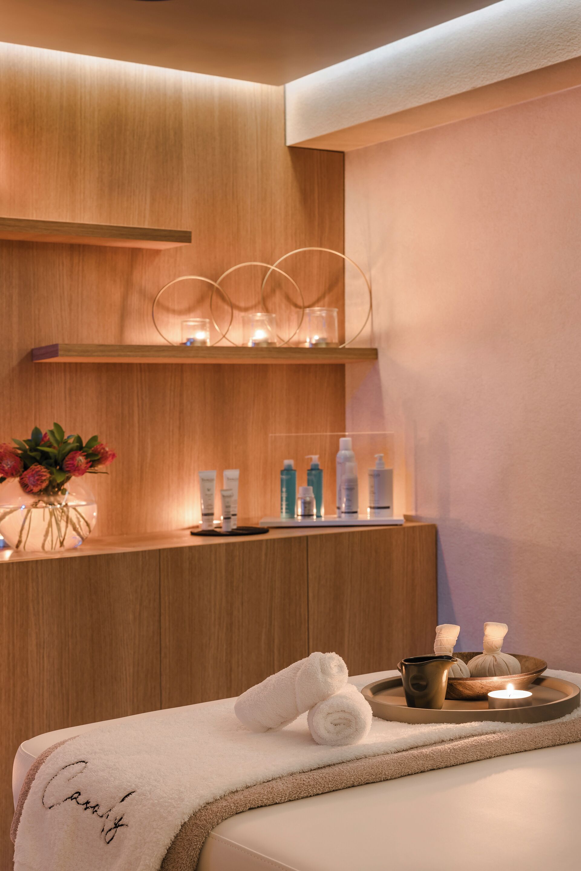 A modern spa room with a wooden reception desk, a vase of flowers, and a tray with towels and candles.