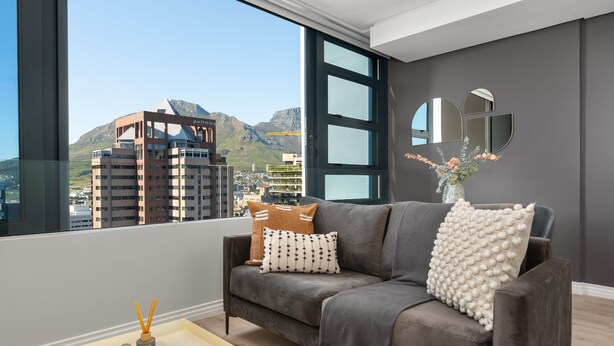 A modern living room with a grey sofa, a coffee table, and a view of a cityscape and mountains. - Modern 16 On Bree Apartment , No Loadshedding (Cape Town)