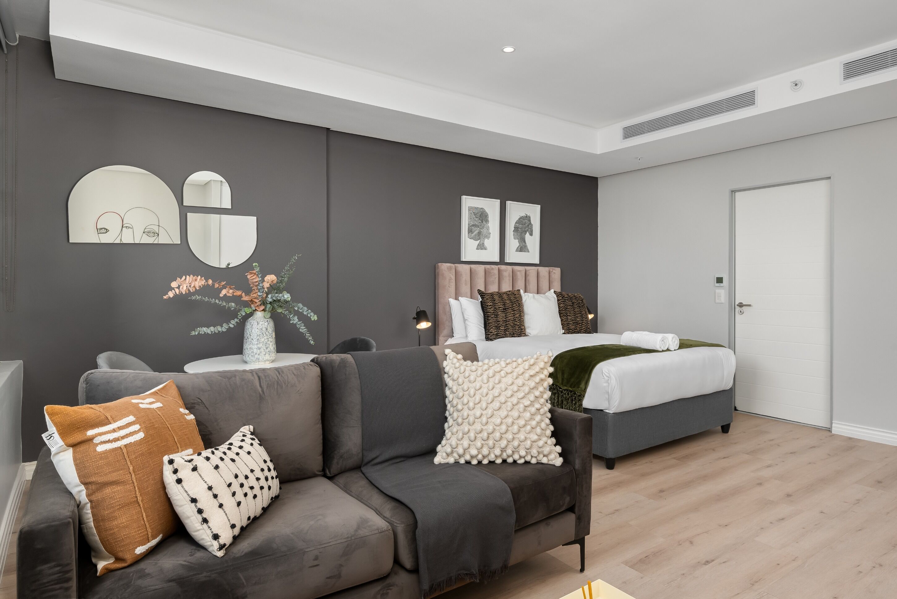 A modern bedroom with a grey sofa, a bed with white bedding, and a grey headboard. There are two wall-mounted mirrors and a vase with flowers on a small table.
