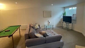 Una sala de estar con mesa de billar, un sofá, un televisor sobre un mueble bajo y una ventana con persianas.