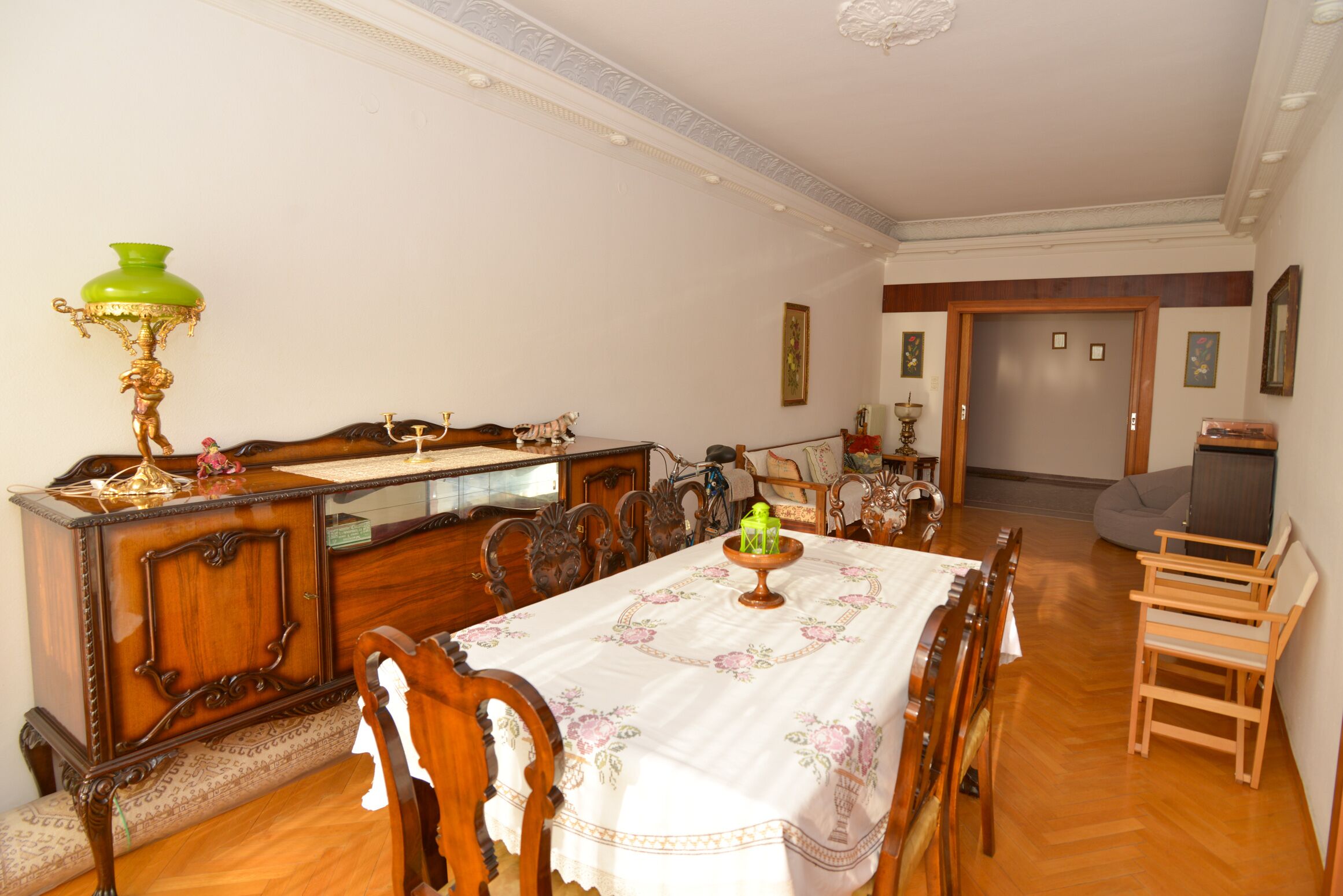 A dining area with a wooden table, chairs, a green lamp, and a television in the background.