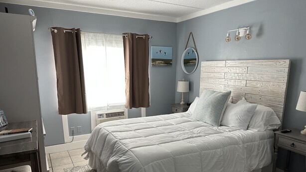 A bedroom with a large bed, a nightstand with a lamp, a dresser, and a window with curtains. - Cute Condo close to Coligny Beach (Hilton Head Island)