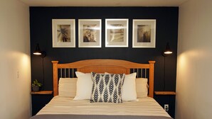 Ein ordentlich bezogenes Bett mit Holz-Kopfstuebe, ein Nachttisch mit Lampe und vier gerahmte Bilder an der Wand ueber dem Bett.