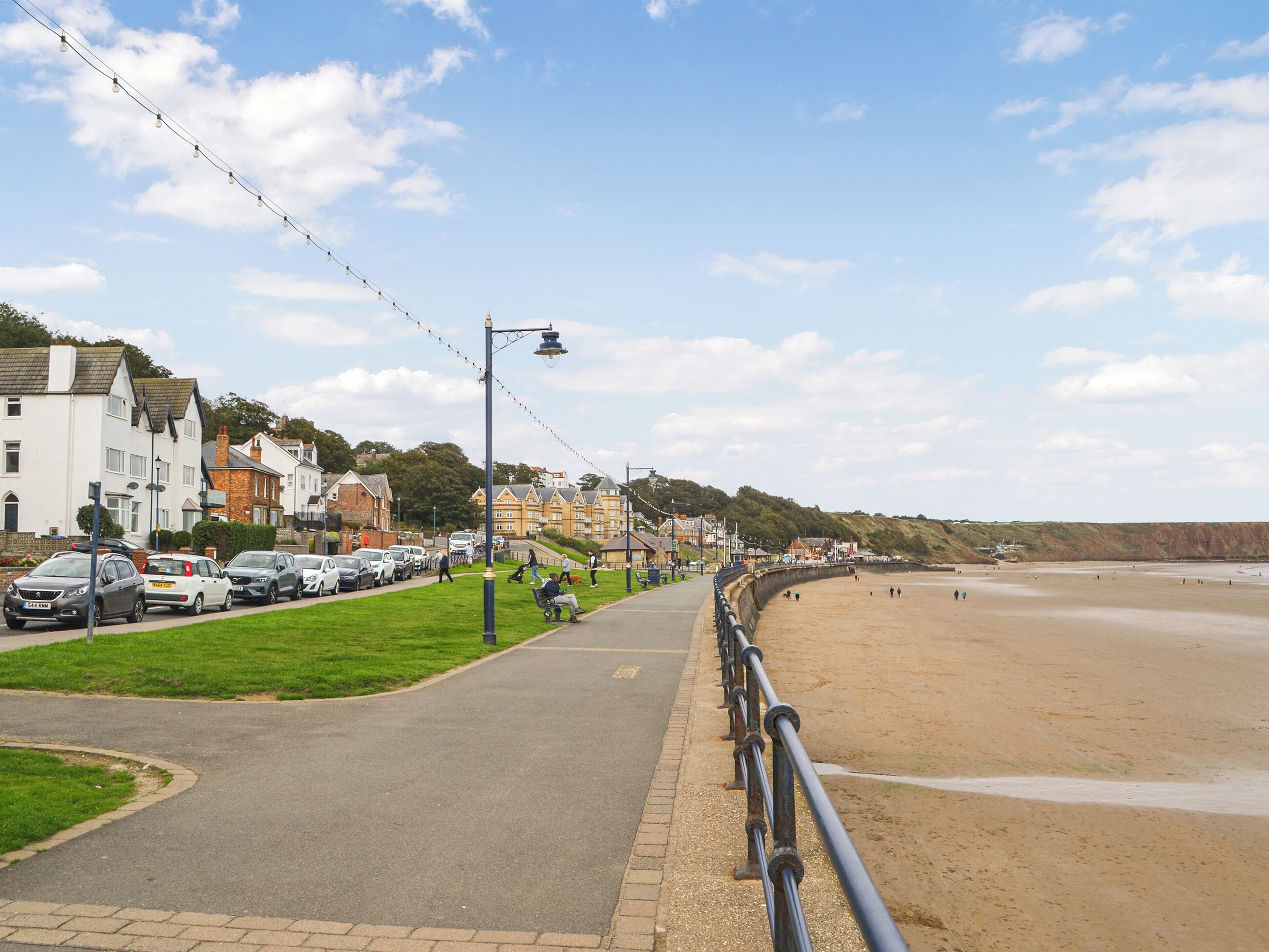 A coastal promenade with a paved walkway, parked cars, and buildings in the background.