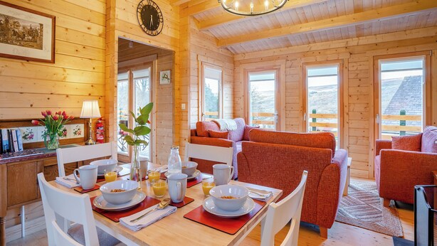 A wooden cabin interior with a dining table set for breakfast, a sofa, and a view of the outdoors. - 2 bedroom accommodation in Garnant Ammanford (Garnant Ammanford)