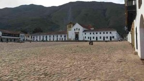 A courtyard with cobblestone flooring, surrounded by white buildings with multiple windows and a central building with a bell tower. - Hotel Esquina Colonial (Villa de Leyva)
