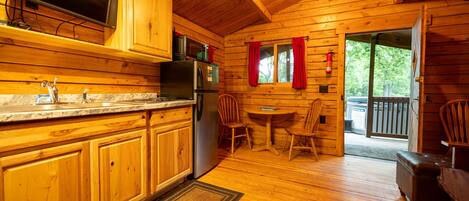 A wooden cabin interior with a kitchen, dining area, and a view through a door.