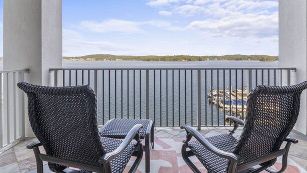 A balcony with two wicker chairs facing a lake, a railing, and a view of a dock and boats.