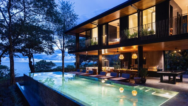 A modern house with a large glass infinity pool, outdoor seating, and a view of the sea.