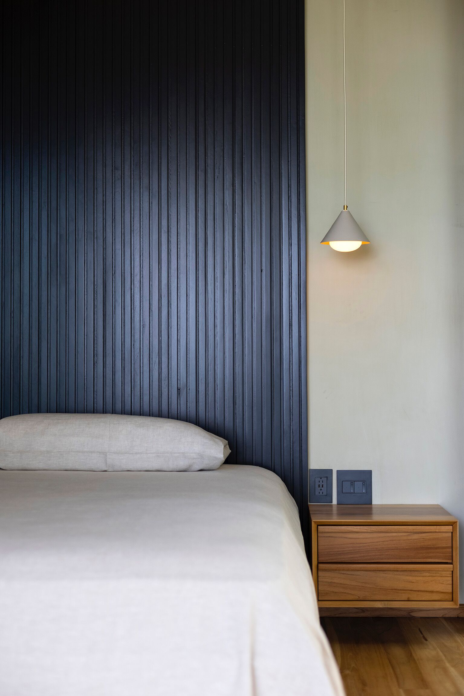 A bedroom with a bed, a nightstand, a wall-mounted lamp, and a power outlet.