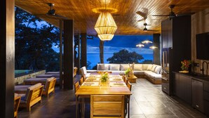A modern outdoor dining area with wooden furniture, a central table set for a meal, and a view of the sky and trees.