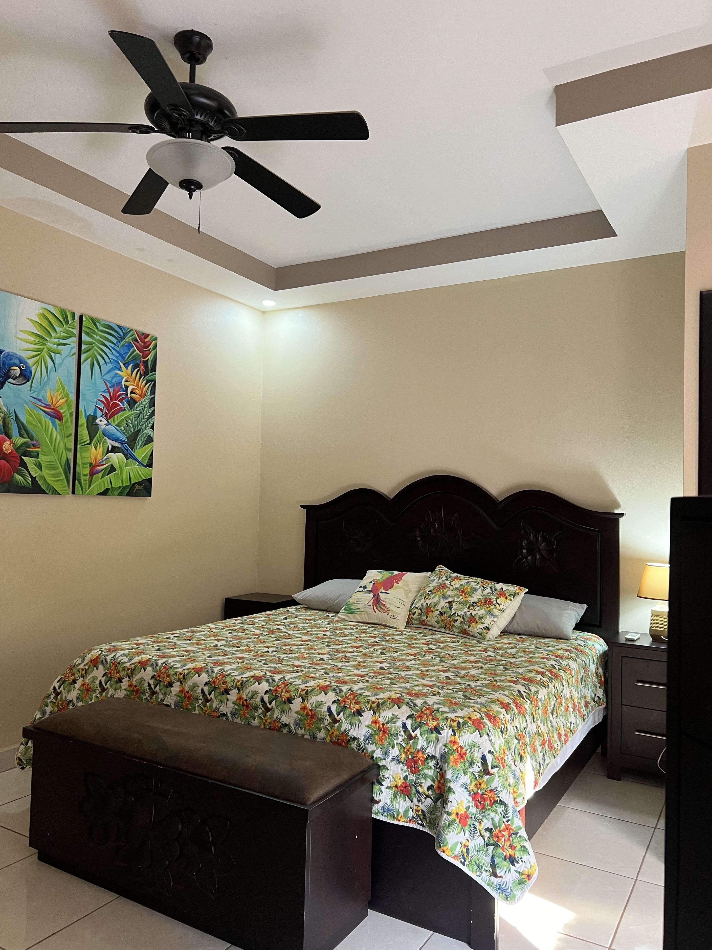 Un dormitorio con cama, un banco, un ventilador de techo, una mesita de noche y un cuadro en la pared.