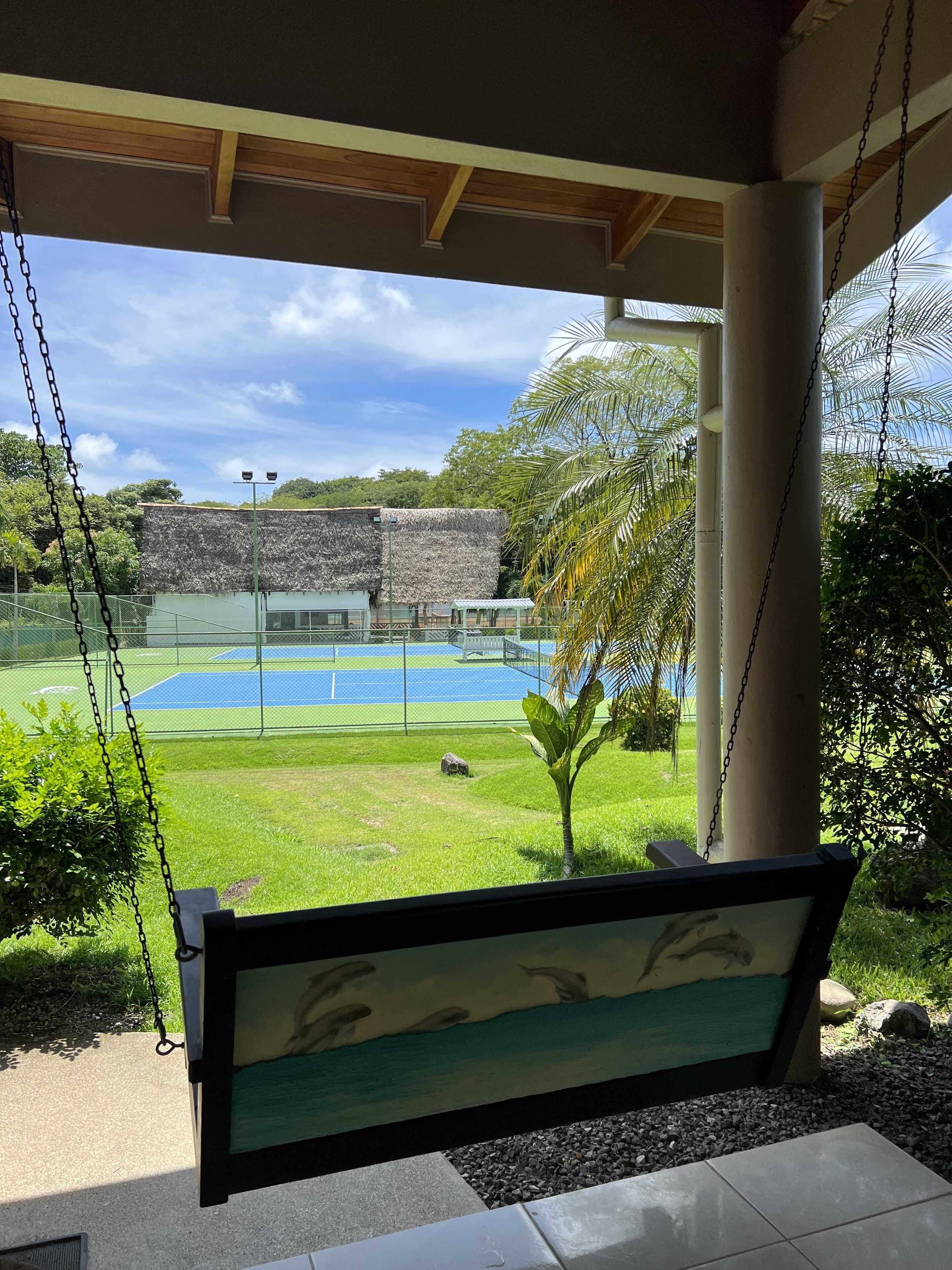 A swing chair overlooking a tennis court and a building with a thatched roof.