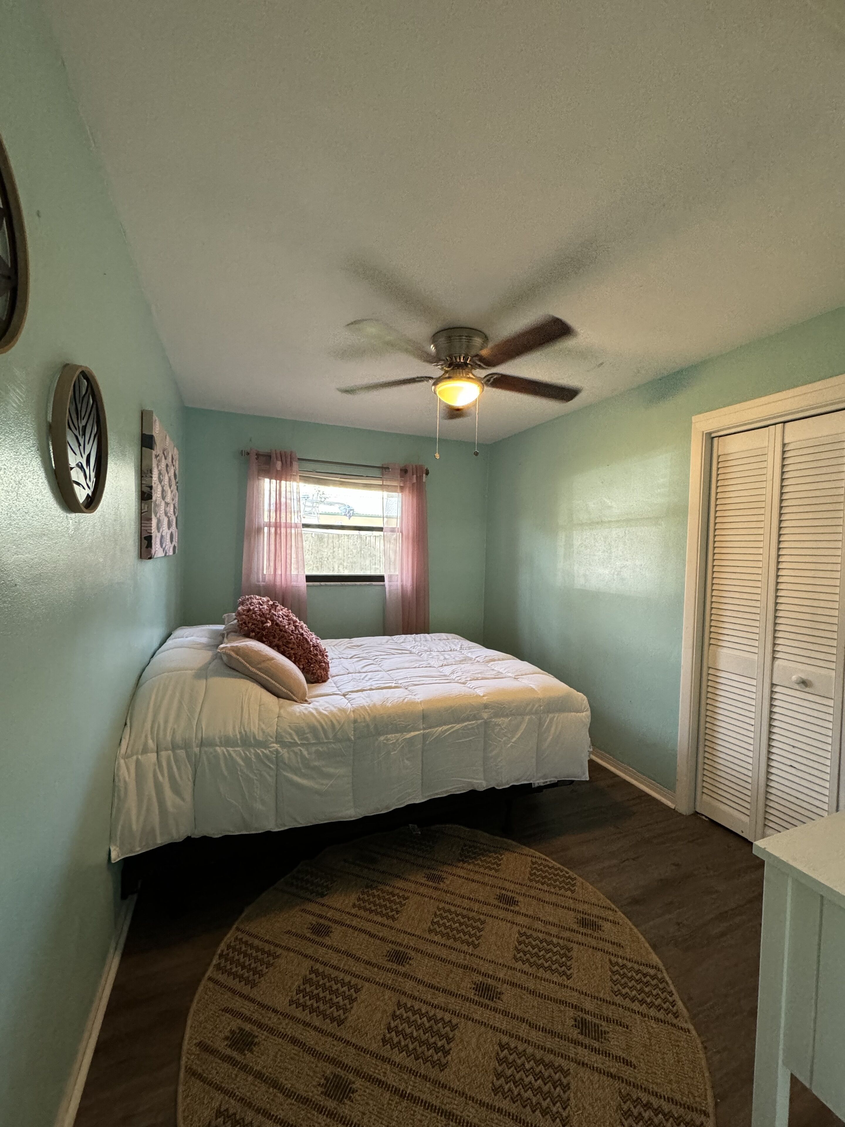 A bedroom with a bed, a ceiling fan, a window with curtains, a door, and a patterned rug.