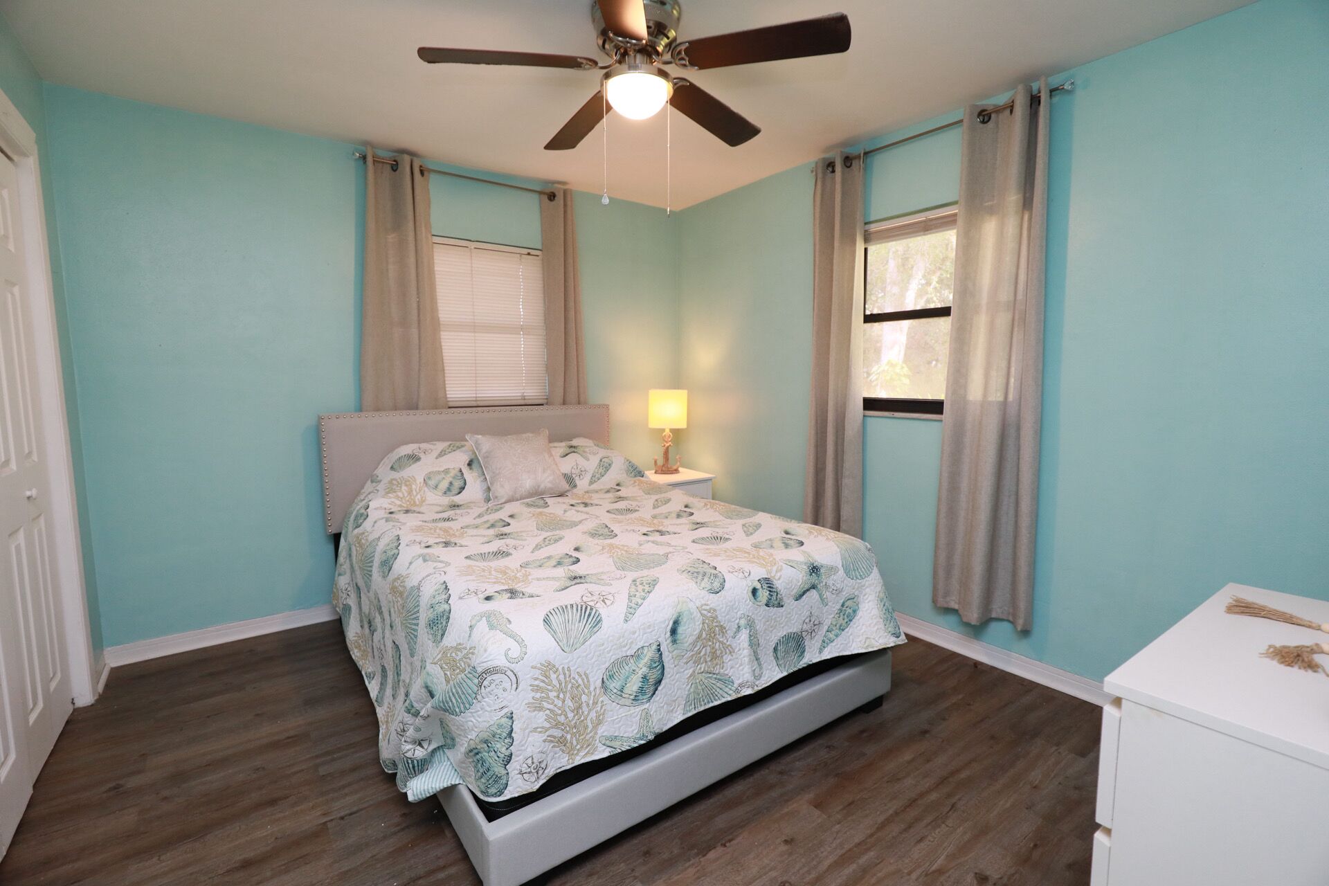 A bedroom with a bed, a ceiling fan, a nightstand with a lamp, and a dresser.