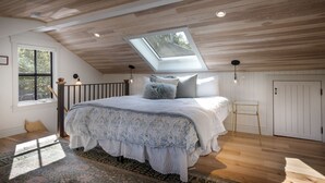 A bedroom with a sloped ceiling, a skylight, a bed with patterned bedding, a small wooden stool, and a view of the outdoors through a window. - Charming Loft Bedroom Cottage in Downtown White, 1 block away from everything (Whitefish)
